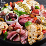 a platter of cold appetizers including ham, prosciutto, salami, blue cheese, mozzarella with pesto and olives