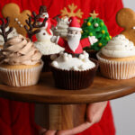 Woman holding tasty Christmas cupcakes on light background