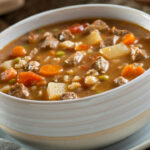 A bowl of delicious beef and barley soup with carrots, tomato, potato, celery, and peas.