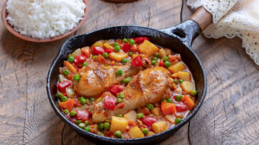 Chicken Afritada. Braised Chicken in Tomato Sauce mixed with potatoes, carrots, bell pepper, and green peas served in frying pan with bowl of cooked rice.