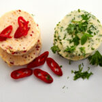Variety of circular flavored butter pats on white viewed top down with fresh chili peppers, lemon, parsley and ground black pepper