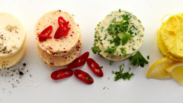 Variety of circular flavored butter pats on white viewed top down with fresh chili peppers, lemon, parsley and ground black pepper