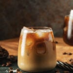 Ice coffee in a tall glass with cream poured over, ice cubes and beans on a dark concrete table.