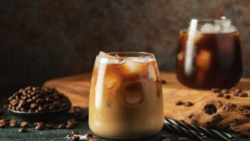 Ice coffee in a tall glass with cream poured over, ice cubes and beans on a dark concrete table.