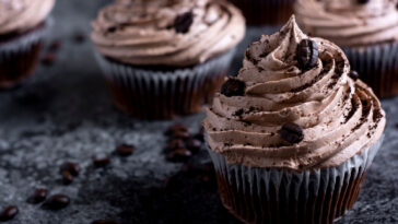Mocha cupcakes topped with ground coffee and coffee beans. Dark food background with chocolate, coffee beans and whisk