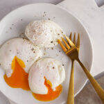 Three poached eggs with egg yolk on a white plate on a marble board and golden forks