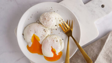 Three poached eggs with egg yolk on a white plate on a marble board and golden forks