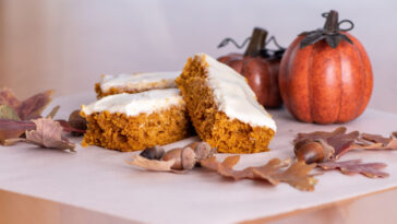 Pumpkin bars topped with cream cheese frosting surrounded with leaves, acorns, pumpkins and fall decor.