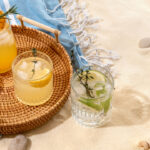 Selection of summer alcoholic cocktails on a wicker tray on beach with white sand.