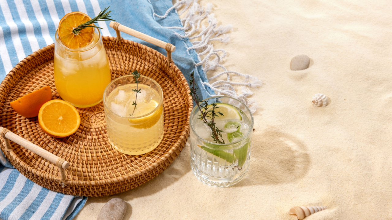 Selection of summer alcoholic cocktails on a wicker tray on beach with white sand.