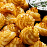close-up of Duchess potatoes, mashed potatoes piped in decorative swirls, browned in an oven served with white sauce on a black plate on a wooden table