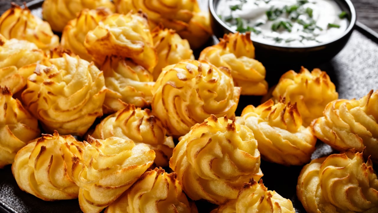 close-up of Duchess potatoes, mashed potatoes piped in decorative swirls, browned in an oven served with white sauce on a black plate on a wooden table