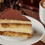 A slice of tiramisu cake on a plate with a cappuccino on a wooden table